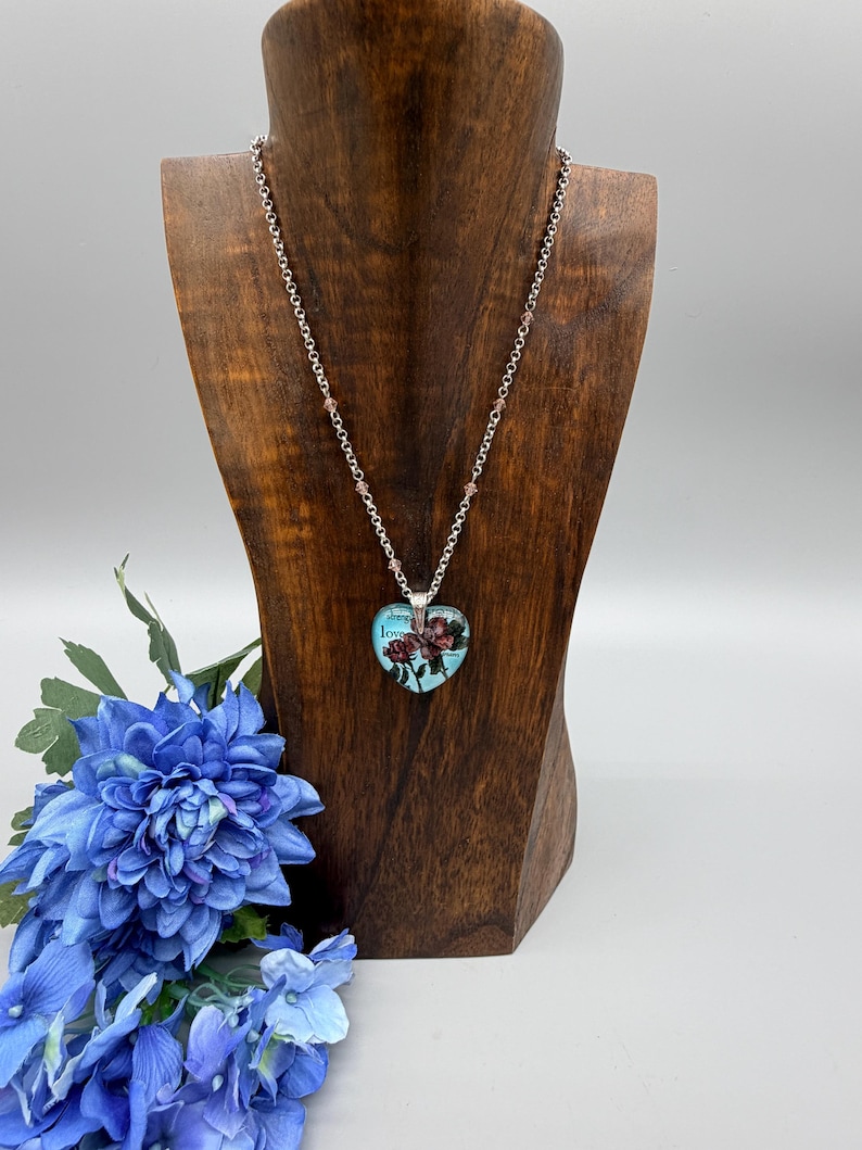 May include: A silver-toned necklace with a heart-shaped pendant. The pendant features a floral design with the word "love" and is displayed on a wooden jewelry stand. Blue flowers are in the foreground.