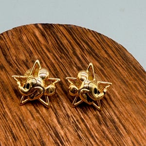 May include: A pair of gold-colored star-shaped stud earrings. Each earring features a Mickey Mouse design in the center. The earrings are displayed on a wooden surface with a natural wood grain pattern.