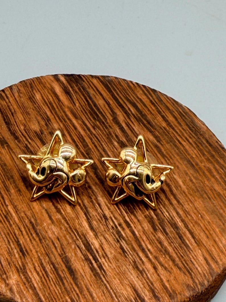 May include: A pair of gold-colored star-shaped stud earrings. Each earring features a Mickey Mouse design in the center. The earrings are displayed on a wooden surface with a natural wood grain pattern.