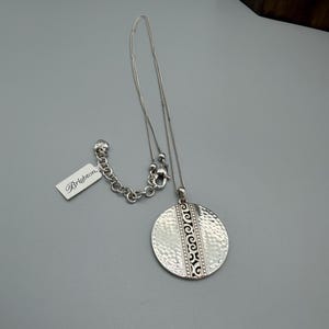 Puede incluir: Un collar Brighton plateado con un colgante texturizado y martillado. El colgante redondo presenta un dise&ntilde;o grabado y cuelga de una delicada cadena. Una etiqueta con el logotipo de Brighton est&aacute; adjunta. El collar se muestra sobre una superficie gris.
