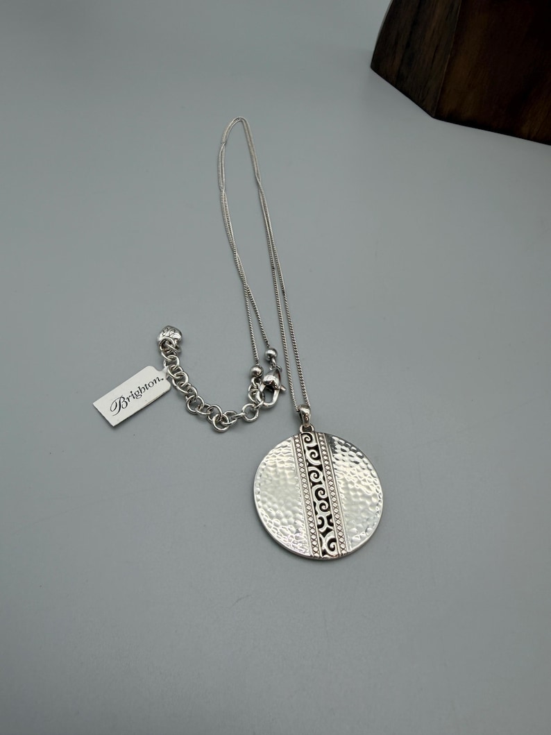 Puede incluir: Un collar Brighton plateado con un colgante texturizado y martillado. El colgante redondo presenta un dise&ntilde;o grabado y cuelga de una delicada cadena. Una etiqueta con el logotipo de Brighton est&aacute; adjunta. El collar se muestra sobre una superficie gris.