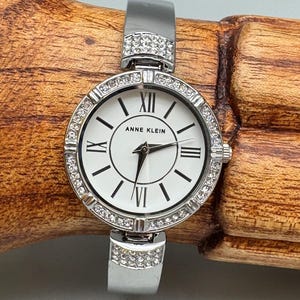 Può includere: Orologio Anne Klein argentato con quadrante bianco e numeri romani. L'orologio presenta una lunetta e dettagli del cinturino tempestati di cristalli. L'orologio ha un cinturino argentato e un quadrante rotondo. Un accessorio alla moda.
