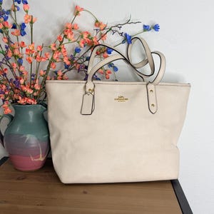 May include: A cream-colored tote bag with two shoulder straps, gold-tone hardware, and a hanging leather luggage tag. The front features the gold Coach logo. The bag has a structured rectangular shape with a textured finish and reinforced strap attachments.