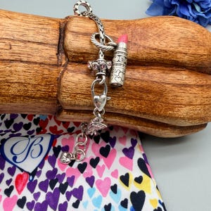 May include: A silver charm bracelet featuring a lipstick charm with a pink tip, a heart-shaped clasp, and a spacer bead with small pink stones. The bracelet has a braided chain design and includes a small heart charm at the end of the extender chain.
