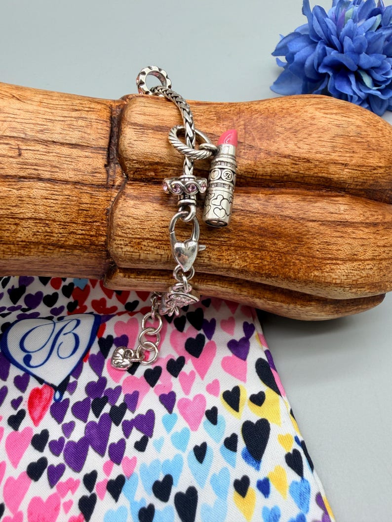 May include: A silver charm bracelet featuring a lipstick charm with a pink tip, a heart-shaped clasp, and a spacer bead with small pink stones. The bracelet has a braided chain design and includes a small heart charm at the end of the extender chain.