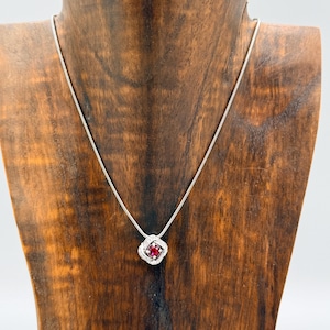 May include: A silver necklace with a square pendant featuring a red gemstone surrounded by small white stones. The pendant is suspended from a delicate silver chain. The necklace is displayed on a wooden jewelry stand.