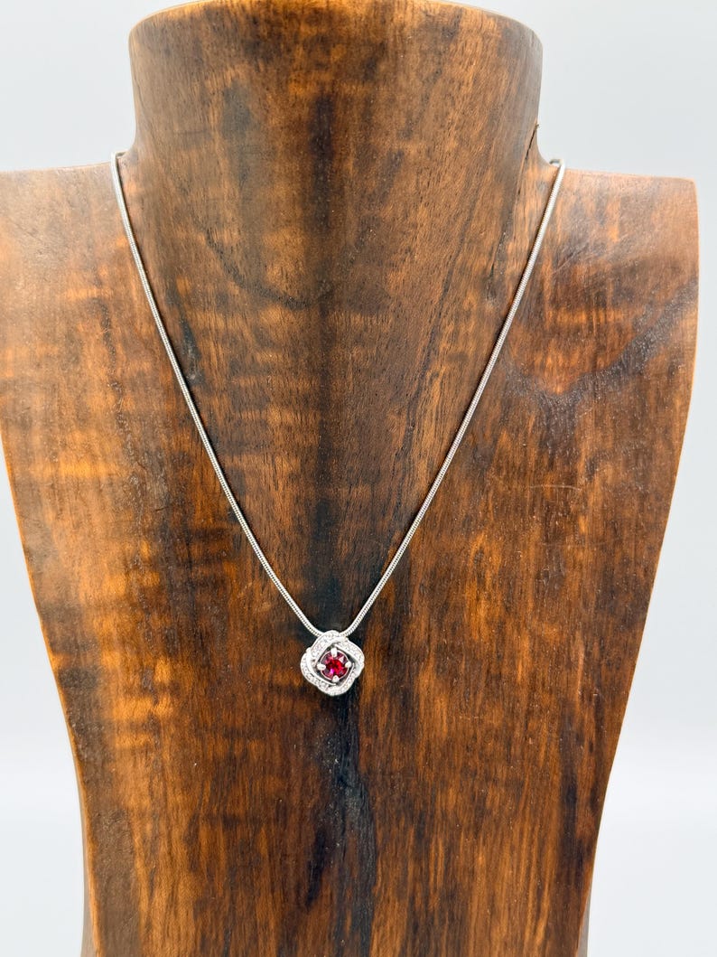 May include: A silver necklace with a square pendant featuring a red gemstone surrounded by small white stones. The pendant is suspended from a delicate silver chain. The necklace is displayed on a wooden jewelry stand.