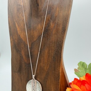 Puede incluir: Collar con colgante plateado con un colgante circular texturizado. El colgante tiene un acabado martillado y un dise&ntilde;o vertical. El collar se exhibe en un expositor de madera, con una flor naranja en el fondo.