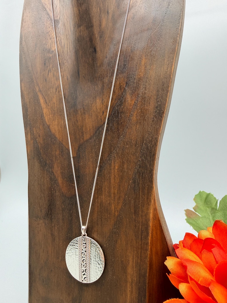 Puede incluir: Collar con colgante plateado con un colgante circular texturizado. El colgante tiene un acabado martillado y un dise&ntilde;o vertical. El collar se exhibe en un expositor de madera, con una flor naranja en el fondo.