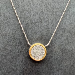 Puede incluir: Un collar plateado con un colgante circular dorado. El colgante est&aacute; incrustado con peque&ntilde;os cristales transparentes. El collar se muestra sobre una superficie oscura y texturizada. El colgante mide aproximadamente 2,5 cm de di&aacute;metro.
