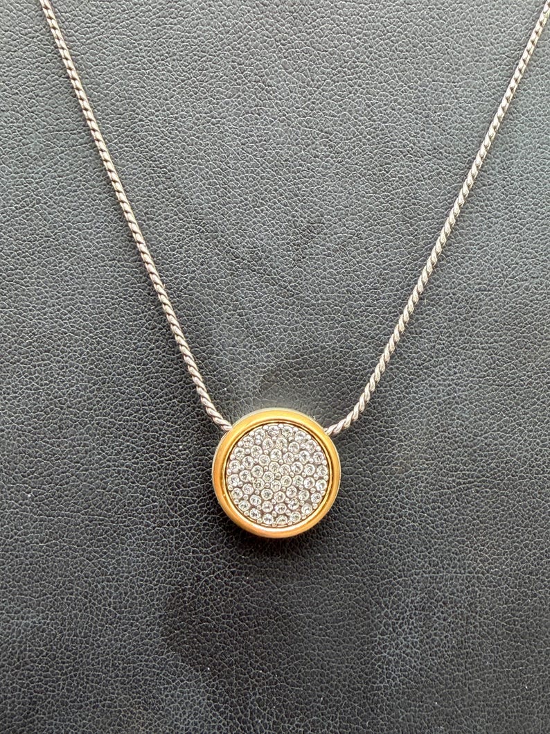 Puede incluir: Un collar plateado con un colgante circular dorado. El colgante est&aacute; incrustado con peque&ntilde;os cristales transparentes. El collar se muestra sobre una superficie oscura y texturizada. El colgante mide aproximadamente 2,5 cm de di&aacute;metro.