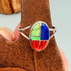 May include: A silver ring featuring an oval inlay design composed of four distinct segments: a light green stone, a vibrant blue stone, a small iridescent opal-like piece, and a textured red stone at the base, set in a polished metal band.