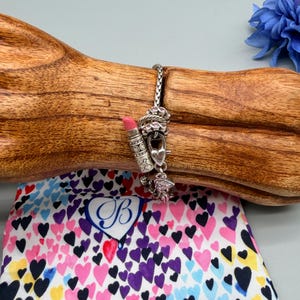 May include: A silver chain bracelet featuring multiple charms, including a lipstick tube with a pink tip, a heart-shaped padlock, and small round charms with pink accents. The bracelet is displayed on a wooden form above a fabric pouch with hearts.
