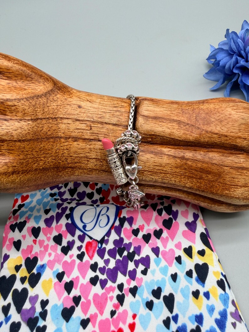 May include: A silver chain bracelet featuring multiple charms, including a lipstick tube with a pink tip, a heart-shaped padlock, and small round charms with pink accents. The bracelet is displayed on a wooden form above a fabric pouch with hearts.