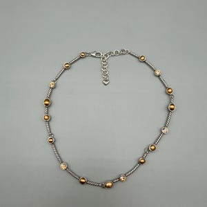 May include: A silver-toned necklace featuring a twisted cable chain design. The chain is accented with alternating round, metallic gold-colored beads and small, clear, faceted crystals. It includes a lobster claw clasp and an adjustable chain extender.