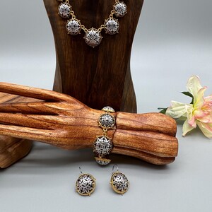 May include: A gold-toned necklace, bracelet, and earrings set. The jewelry features silver-toned, circular pendants with an intricate, symmetrical design. The set is displayed on a wooden jewelry stand and a wooden arm, with a flower in the background.