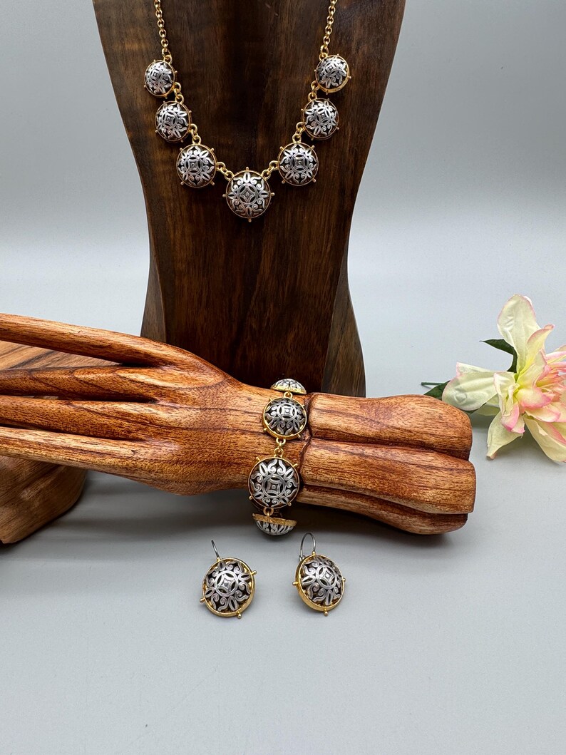 May include: A gold-toned necklace, bracelet, and earrings set. The jewelry features silver-toned, circular pendants with an intricate, symmetrical design. The set is displayed on a wooden jewelry stand and a wooden arm, with a flower in the background.