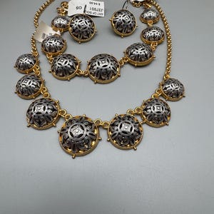 May include: A gold-toned necklace, earrings, and bracelet set. The jewelry features silver and gold circular pendants with an ornate design. The set includes a necklace, a pair of earrings, and a bracelet. The tags read "ART OF GOLD".