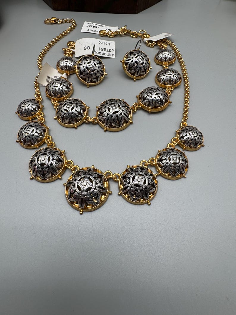 May include: A gold-toned necklace, earrings, and bracelet set. The jewelry features silver and gold circular pendants with an ornate design. The set includes a necklace, a pair of earrings, and a bracelet. The tags read "ART OF GOLD".