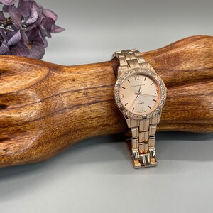 May include: A gold-tone watch with a round face and a gold-tone metal band. The watch face has a white background with black Roman numeral hour markers and a small seconds dial at the 6 o'clock position. The watch face also has a small date window at the 3 o'clock position. The watch is set on a wooden surface.