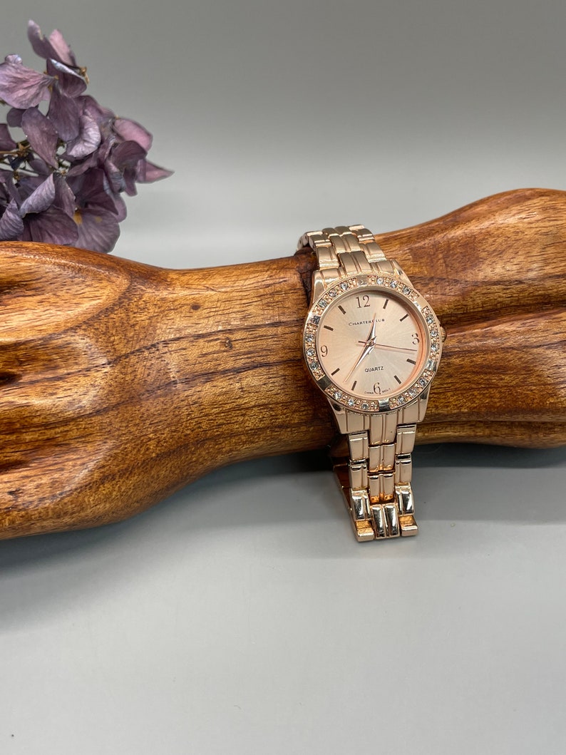 May include: A gold-tone watch with a round face and a gold-tone metal band. The watch face has a white background with black Roman numeral hour markers and a small seconds dial at the 6 o'clock position. The watch face also has a small date window at the 3 o'clock position. The watch is set on a wooden surface.