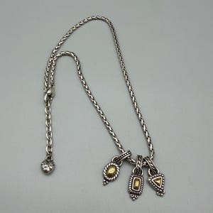 May include: A silver-toned braided chain necklace featuring three dangling charms, each with a central gold-toned accent. The charms are oval, rectangular, and triangular in shape. The necklace includes a lobster claw clasp and a heart-shaped charm.