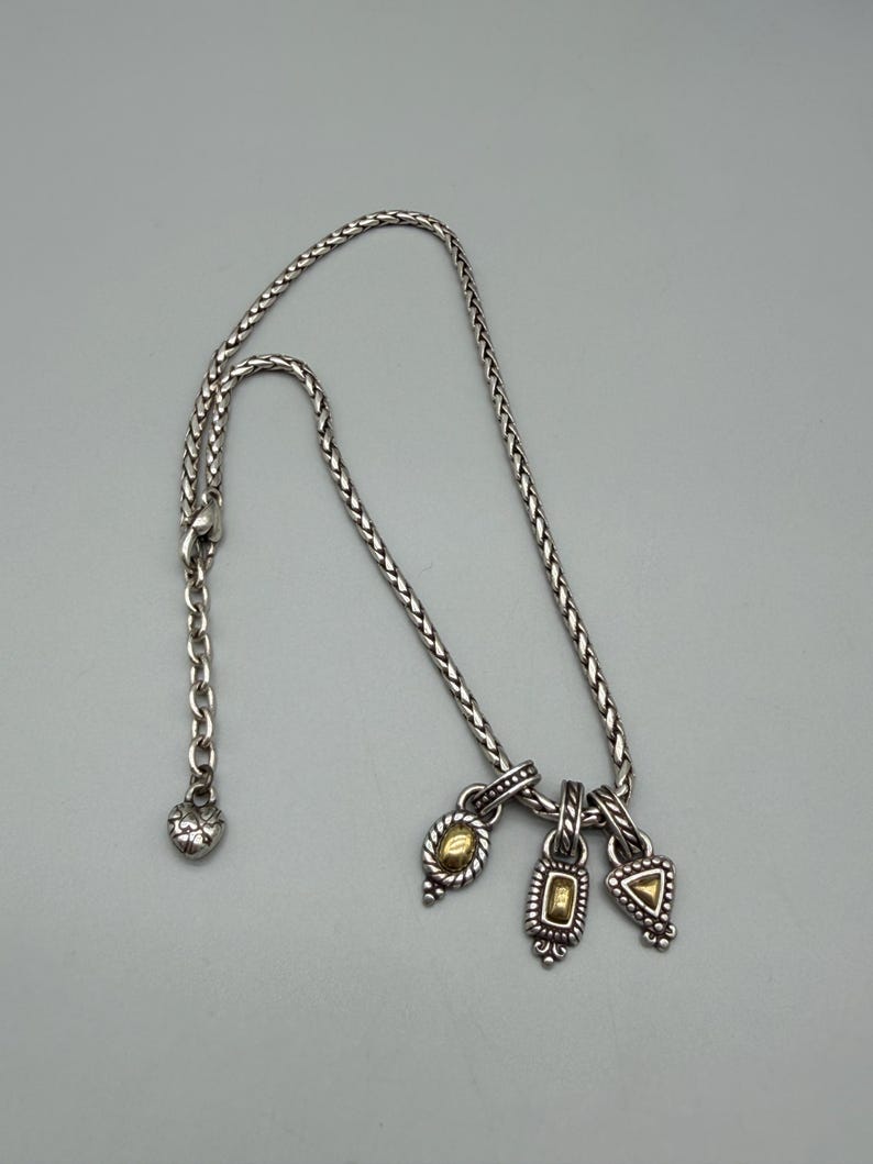 May include: A silver-toned braided chain necklace featuring three dangling charms, each with a central gold-toned accent. The charms are oval, rectangular, and triangular in shape. The necklace includes a lobster claw clasp and a heart-shaped charm.
