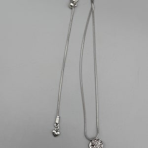 May include: A silver-toned necklace with two pendants. One pendant is a small heart, and the other is a round, textured design with a crisscross pattern. The necklace has a delicate chain and a clasp.