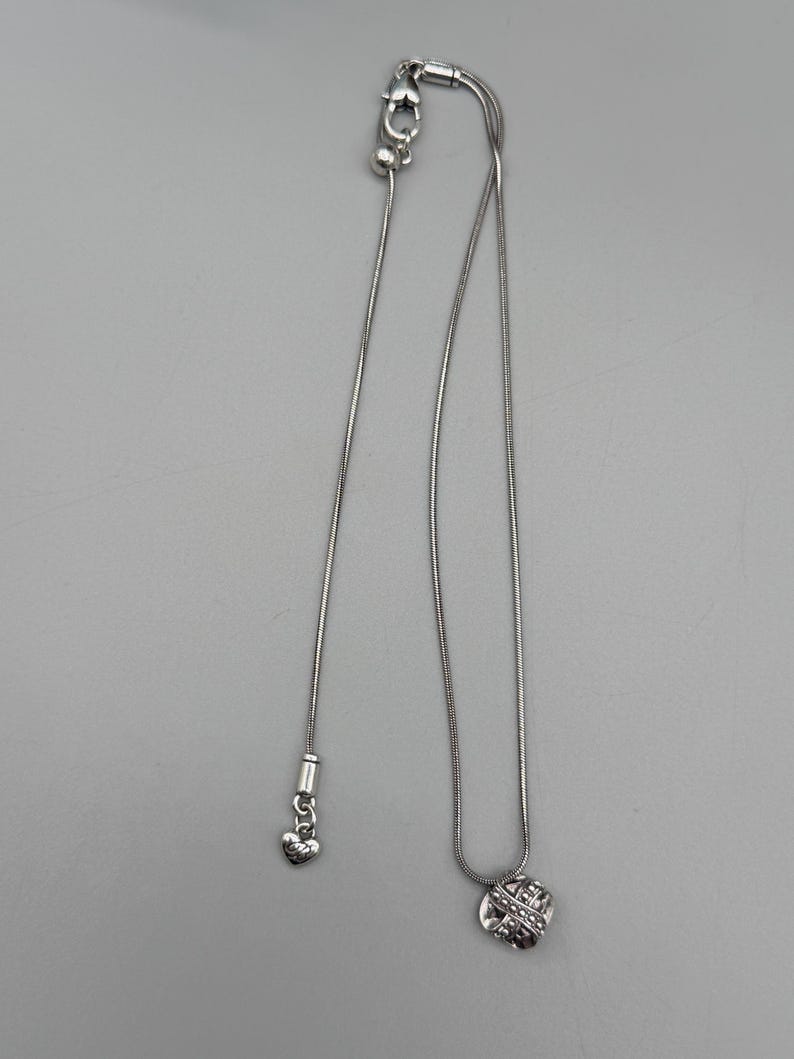 May include: A silver-toned necklace with two pendants. One pendant is a small heart, and the other is a round, textured design with a crisscross pattern. The necklace has a delicate chain and a clasp.