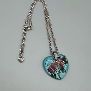 May include: Silver-toned necklace with a heart-shaped pendant. The pendant is light blue with a floral design and the words "strength", "love", "hope", and "dream". The necklace has a heart-shaped charm near the clasp.