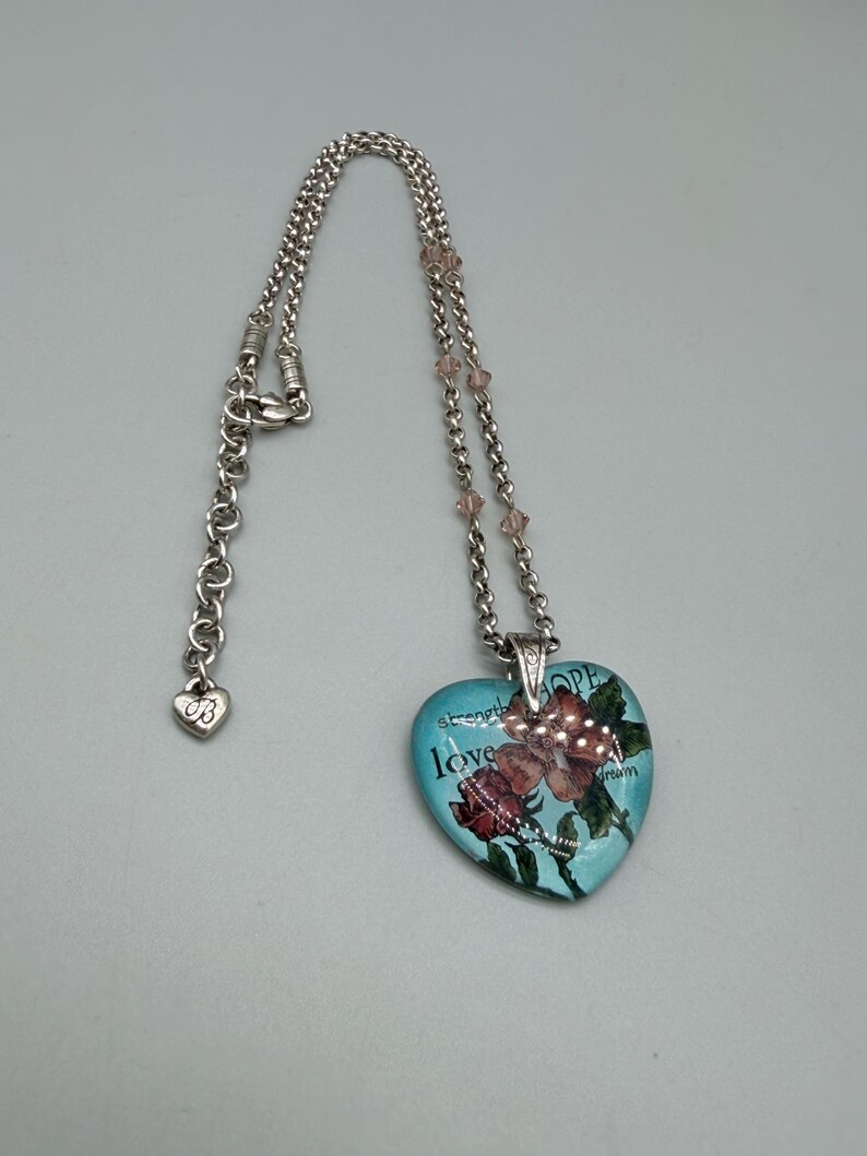 May include: Silver-toned necklace with a heart-shaped pendant. The pendant is light blue with a floral design and the words "strength", "love", "hope", and "dream". The necklace has a heart-shaped charm near the clasp.