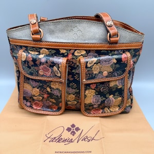 May include: A floral print tote bag with two front pockets. The bag features brown leather handles and trim, with a gray suede-like top. The bag is sitting on a tan bag with the text "Patricia Nash" and "patricianashdesigns.com".