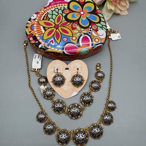 May include: A gold-toned jewelry set with silver and black accents, including a necklace, bracelet, and earrings. The set is displayed with a heart-shaped tin featuring a colorful floral design. A tag is attached to the jewelry.