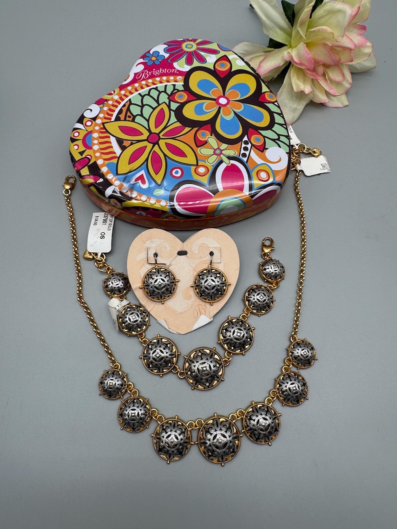 May include: A gold-toned jewelry set with silver and black accents, including a necklace, bracelet, and earrings. The set is displayed with a heart-shaped tin featuring a colorful floral design. A tag is attached to the jewelry.