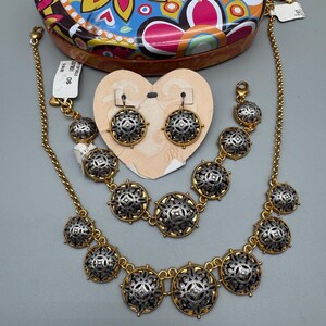May include: A gold-toned jewelry set featuring a necklace, bracelet, and earrings. Each piece has silver-toned, circular medallions with an ornate design. A colorful floral patterned tin is in the background.