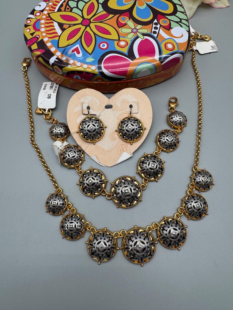 May include: A gold-toned jewelry set featuring a necklace, bracelet, and earrings. Each piece has silver-toned, circular medallions with an ornate design. A colorful floral patterned tin is in the background.