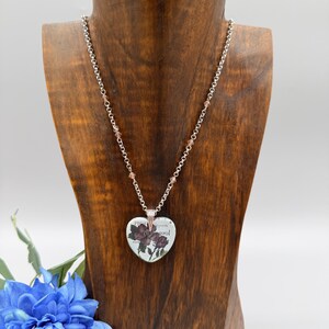 May include: A silver-toned necklace with a heart-shaped pendant featuring a floral design and text. The pendant hangs from a chain with small, pink beads. The necklace is displayed on a wooden bust, with a blue flower in the foreground.