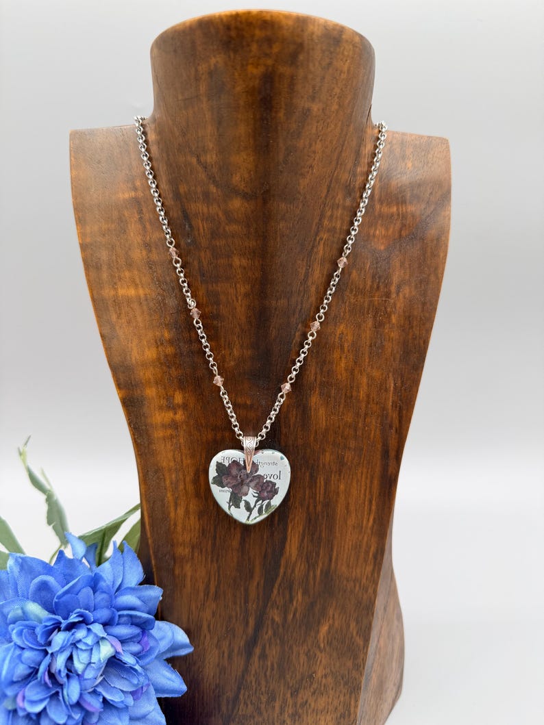 May include: A silver-toned necklace with a heart-shaped pendant featuring a floral design and text. The pendant hangs from a chain with small, pink beads. The necklace is displayed on a wooden bust, with a blue flower in the foreground.