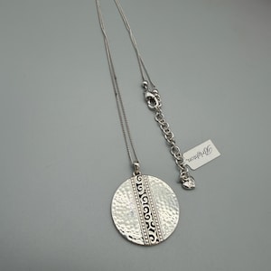 Puede incluir: Collar plateado con un colgante grande y texturizado. El colgante tiene un dise&ntilde;o decorativo central y cuelga de una cadena fina. Una etiqueta con la palabra "Brighton" est&aacute; unida a la cadena.