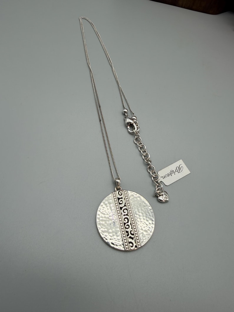 Puede incluir: Collar plateado con un colgante grande y texturizado. El colgante tiene un dise&ntilde;o decorativo central y cuelga de una cadena fina. Una etiqueta con la palabra "Brighton" est&aacute; unida a la cadena.