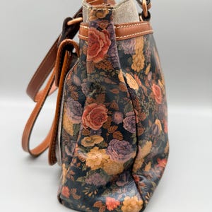 May include: A floral-patterned handbag with a dark background and a variety of colorful roses in shades of red, orange, and purple. The bag has brown leather straps and trim, and a light-colored interior. The bag is a shoulder bag style.