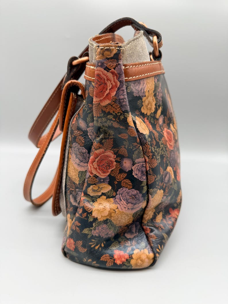 May include: A floral-patterned handbag with a dark background and a variety of colorful roses in shades of red, orange, and purple. The bag has brown leather straps and trim, and a light-colored interior. The bag is a shoulder bag style.