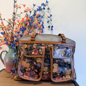 May include: A floral-patterned handbag with two front pockets and brown leather trim. The bag features a dark background with colorful roses and other flowers. The handles and top are brown leather. The bag is sitting on a wooden surface.