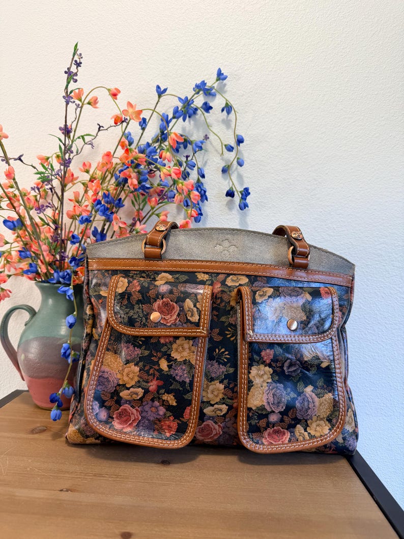 May include: A floral-patterned handbag with two front pockets and brown leather trim. The bag features a dark background with colorful roses and other flowers. The handles and top are brown leather. The bag is sitting on a wooden surface.