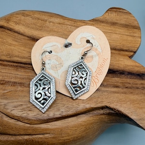 Puede incluir: Un par de pendientes plateados con forma hexagonal. Cada pendiente presenta un dise&ntilde;o de filigrana dentro de un marco de peque&ntilde;os cristales transparentes. Los pendientes se exhiben en una etiqueta en forma de coraz&oacute;n con la palabra "Brighton".