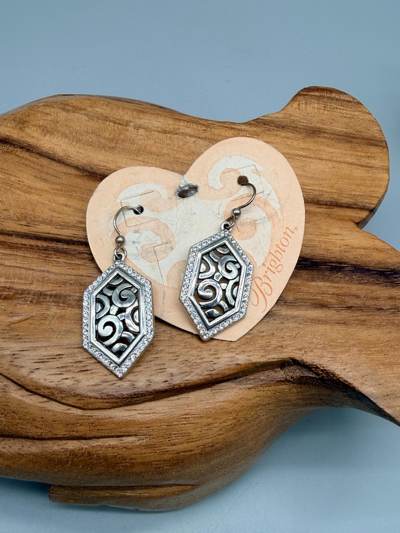 Puede incluir: Un par de pendientes plateados con forma hexagonal. Cada pendiente presenta un dise&ntilde;o de filigrana dentro de un marco de peque&ntilde;os cristales transparentes. Los pendientes se exhiben en una etiqueta en forma de coraz&oacute;n con la palabra "Brighton".