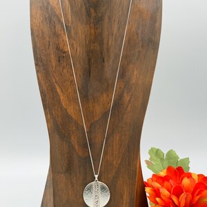 Puede incluir: Un collar con colgante plateado con una cadena larga. El colgante es un c&iacute;rculo texturizado, martillado, con un dise&ntilde;o vertical. El collar se exhibe en un expositor de joyas de madera, con una flor naranja brillante en el fondo.