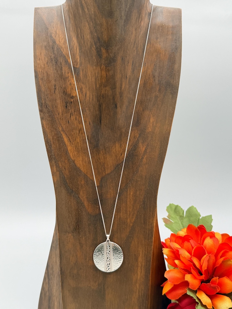 Puede incluir: Un collar con colgante plateado con una cadena larga. El colgante es un c&iacute;rculo texturizado, martillado, con un dise&ntilde;o vertical. El collar se exhibe en un expositor de joyas de madera, con una flor naranja brillante en el fondo.