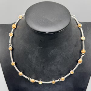 May include: A silver-toned necklace featuring a twisted rope-style chain interspersed with small, circular bezel-set stones. The stones alternate between solid matte gold-colored spheres and iridescent, multi-colored faceted crystals.