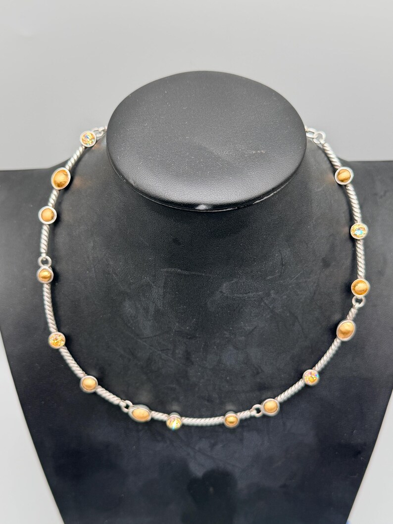May include: A silver-toned necklace featuring a twisted rope-style chain interspersed with small, circular bezel-set stones. The stones alternate between solid matte gold-colored spheres and iridescent, multi-colored faceted crystals.